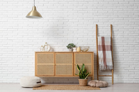 Chest of drawers, ladder with plaid, houseplant and poufs near white brick wall in roomの写真素材