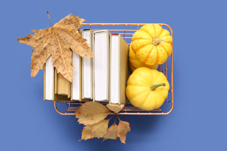 Metal basket with stack of books, pumpkins and autumn leaves on blue backgroundの写真素材