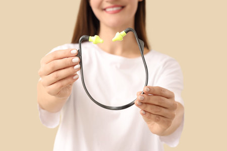 Young woman with ear plugs on beige background, closeup