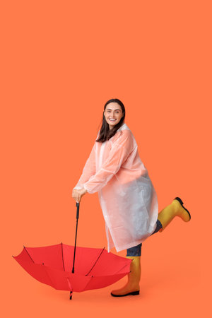 Happy young woman in raincoat and with umbrella on orange backgroundの写真素材