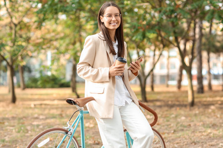 Young businesswoman with bicycle, phone and coffee in city parkの写真素材