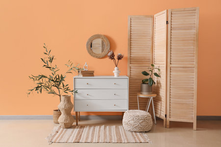 Chest of drawers with houseplants, folding screen and mirror on orange wall in room