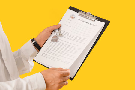 Male real estate agent with key and house rental agreement on yellow background, closeup