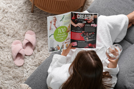 Young woman in bathrobe with glass of water reading magazine on sofa at home, top viewの写真素材