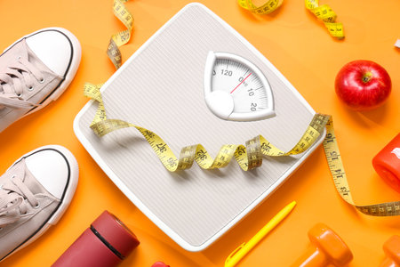 Scales with measuring tape, sport shoes and apple on orange background. Weight loss concept