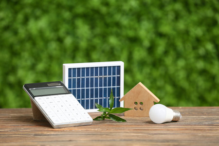 Portable solar panel with house figure, light bulb and calculator on table outdoors. Green Energy...