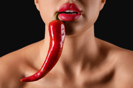 Sexy young woman with chili pepper in mouth on black background, closeupの写真素材