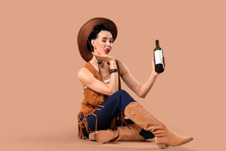 Young pin-up cowgirl with bottle of wine sitting on brown backgroundの写真素材