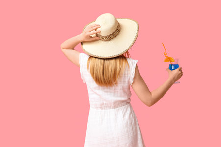 Beautiful young woman in stylish summer outfit with glass of cocktail on pink background, back viewの写真素材