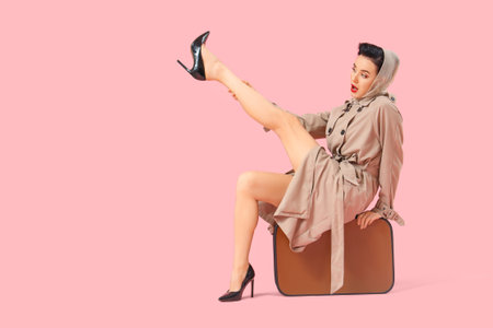 Young pin-up woman in coat with suitcase on pink backgroundの写真素材