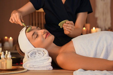 Beautician with facial massage tools and woman lying in dark salonの写真素材