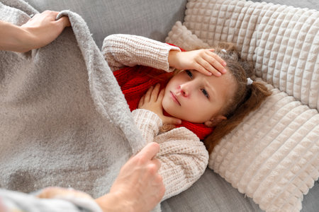 Mother covering with plaid her sick little daughter on sofa at homeの写真素材