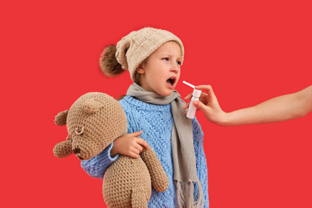 Sick little girl with toy bear and woman spraying her sore throat on red backgroundの写真素材