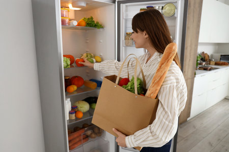 Young woman with bag of food near open refrigerator in kitchenの写真素材