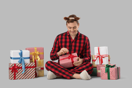 Young man in pajamas with Christmas gifts sitting on gray backgroundの写真素材