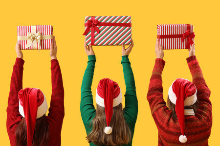 Young women in Santa hats with Christmas gifts on yellow background. Back viewの写真素材