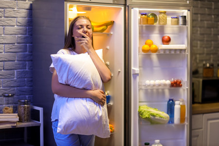 Sleepy young woman with pillow near open refrigerator in kitchen at nightの写真素材