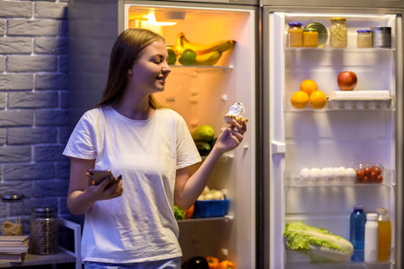 Young woman with mobile phone and cake near open refrigerator in kitchen at nightの写真素材