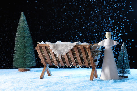 Manger with baby Jesus, angel and Christmas trees on snow against dark background