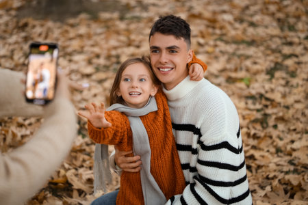 Mother recording video while daughter hugging her happy father in beautiful autumn parkの写真素材