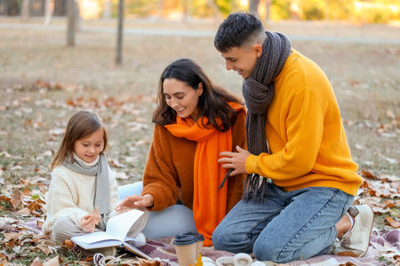 Happy parents with cups of coffee and their cute daughter reading book on picnic in beautiful autumn parkの写真素材