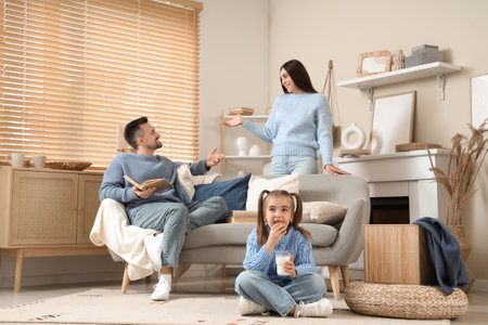 Happy family reading book and their cute little girl in sweater with cup of milk eating cookie at homeの写真素材
