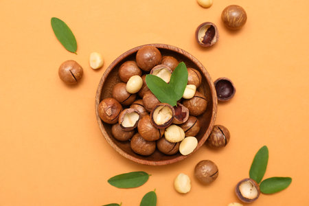 Wooden bowl with tasty macadamia nuts and leaves on orange backgroundの写真素材