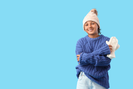 Cute African-American girl in stylish hat and knitted sweater with warm mittens on blue backgroundの写真素材
