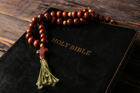 Holy Bible with rosary beads on wooden table, closeup