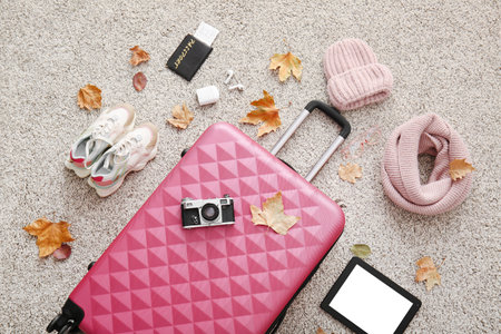 Pink suitcase with photo camera, modern tablet computer, passport, stylish hat, scarf and autumn leaves on gray carpet. Travel conceptの写真素材