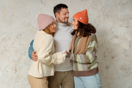 Man and two beautiful women in autumn clothes hugging near white wall. Polyamory concept