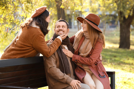 Man with two beautiful women sitting on bench in autumn park. Polyamory concept