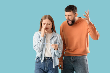 Upset young woman with pregnancy test and her angry husband on blue backgroundの写真素材