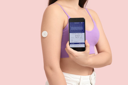 Diabetic young woman with glucose sensor using mobile phone for measuring blood sugar level on pink background, closeupの写真素材