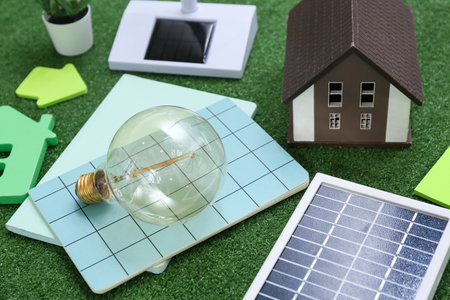 Light bulb with notebooks, house model and solar panel on grass, closeup. Green Energy conceptの写真素材