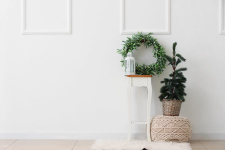Fir tree on pouf and mistletoe wreath with lantern on stand near white wall in room