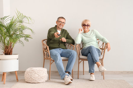 Senior couple sitting in armchairs and watching TV together near white wallの写真素材