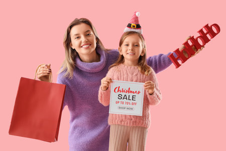 Beautiful young woman with shopping bag and her cute little daughter holding paper with text CHRISTMAS SALE on pink backgroundの写真素材