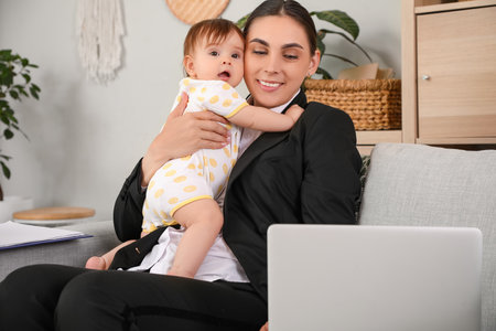 Young happy business mother working with laptop and little baby on sofa at homeの写真素材