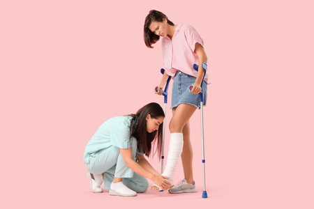 Young woman with broken leg and doctor on pink backgroundの写真素材