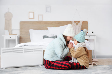 Frozen little girl with her mother holding hands near electric convector heater in bedroomの写真素材