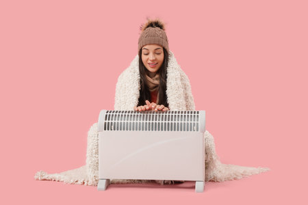 Frozen young woman with plaid and electric convector heater on pink backgroundの写真素材