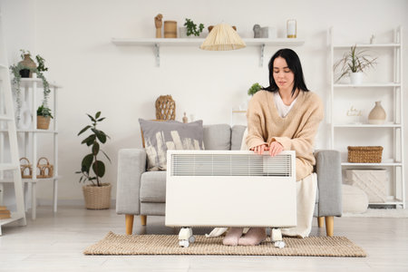 Young woman with electric convector heater sitting on sofa at homeの写真素材