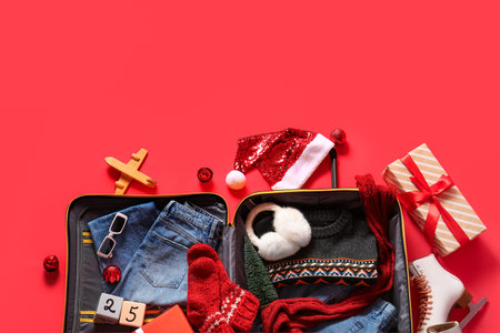 Open suitcase with winter clothes and Christmas decor on red background. Top view