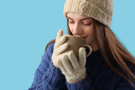 Beautiful young woman in stylish warm clothes with cup of hot tea on blue backgroundの写真素材