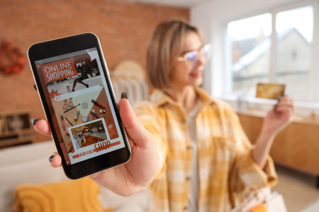 Young woman with mobile phone and credit card shopping online at home on autumn day, closeup