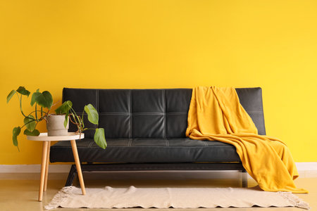 Stylish modern black sofa with plaid and houseplant on table near yellow wallの写真素材