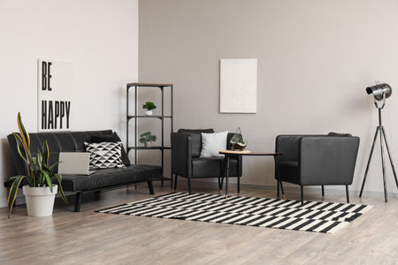 Interior of stylish room with shelving unit, armchairs and laptop on black sofa near white wallの写真素材