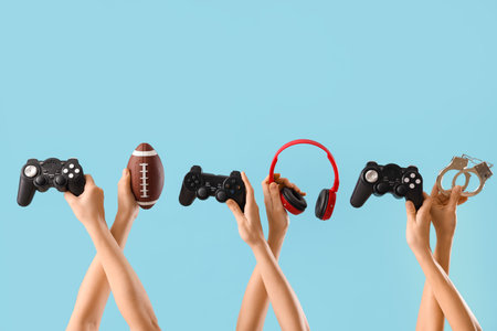 Female hands with game pads, rugby ball, handcuffs and headphones on blue backgroundの写真素材