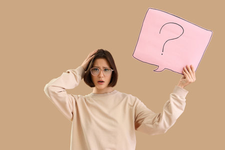 Thoughtful young woman holding speech bubble with question mark on beige backgroundの写真素材